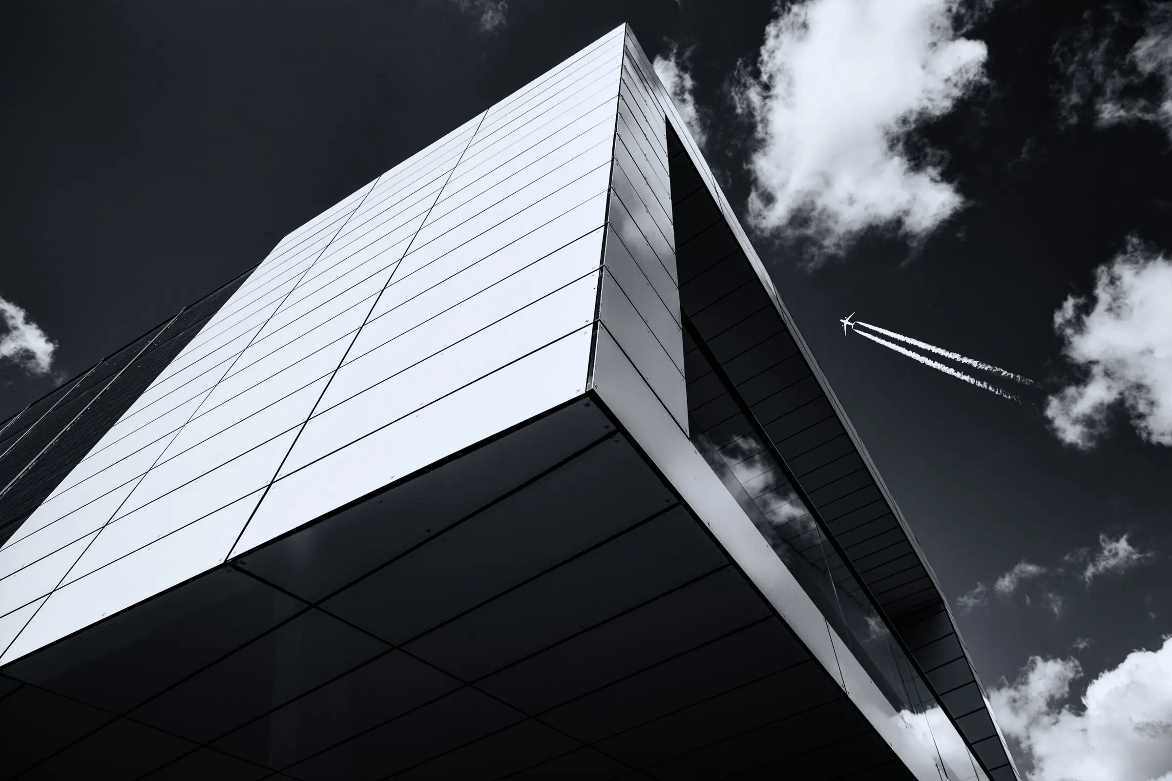 Architekturfotografie in Schwarzweiß: moderne Gebäudeecke mit Metallfassade, darüber ein Flugzeug mit Kondensstreifen
