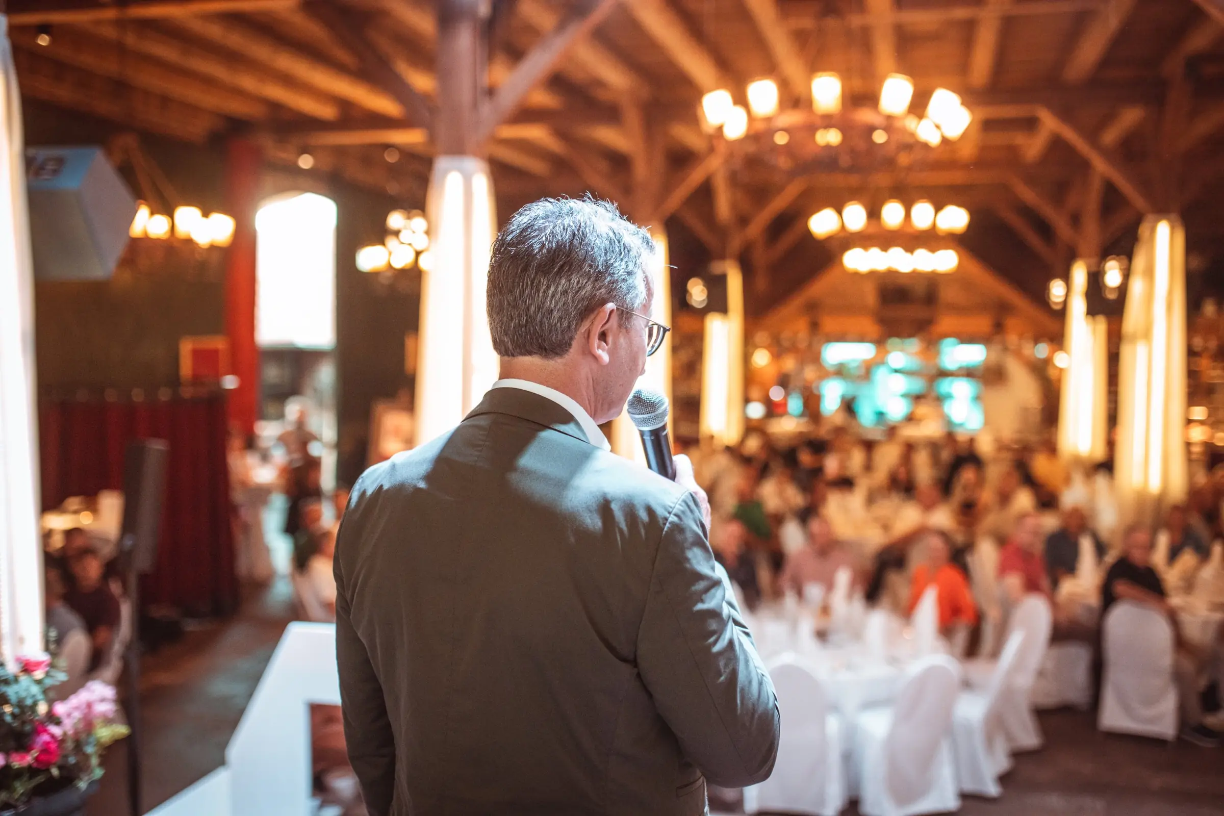 Eventfotografie beim Sommerfest 2026 von HERMANN RIEDE: Redner mit Mikrofon auf der Bühne vor festlich gedeckten Tischen in einer Halle mit Holzdecke