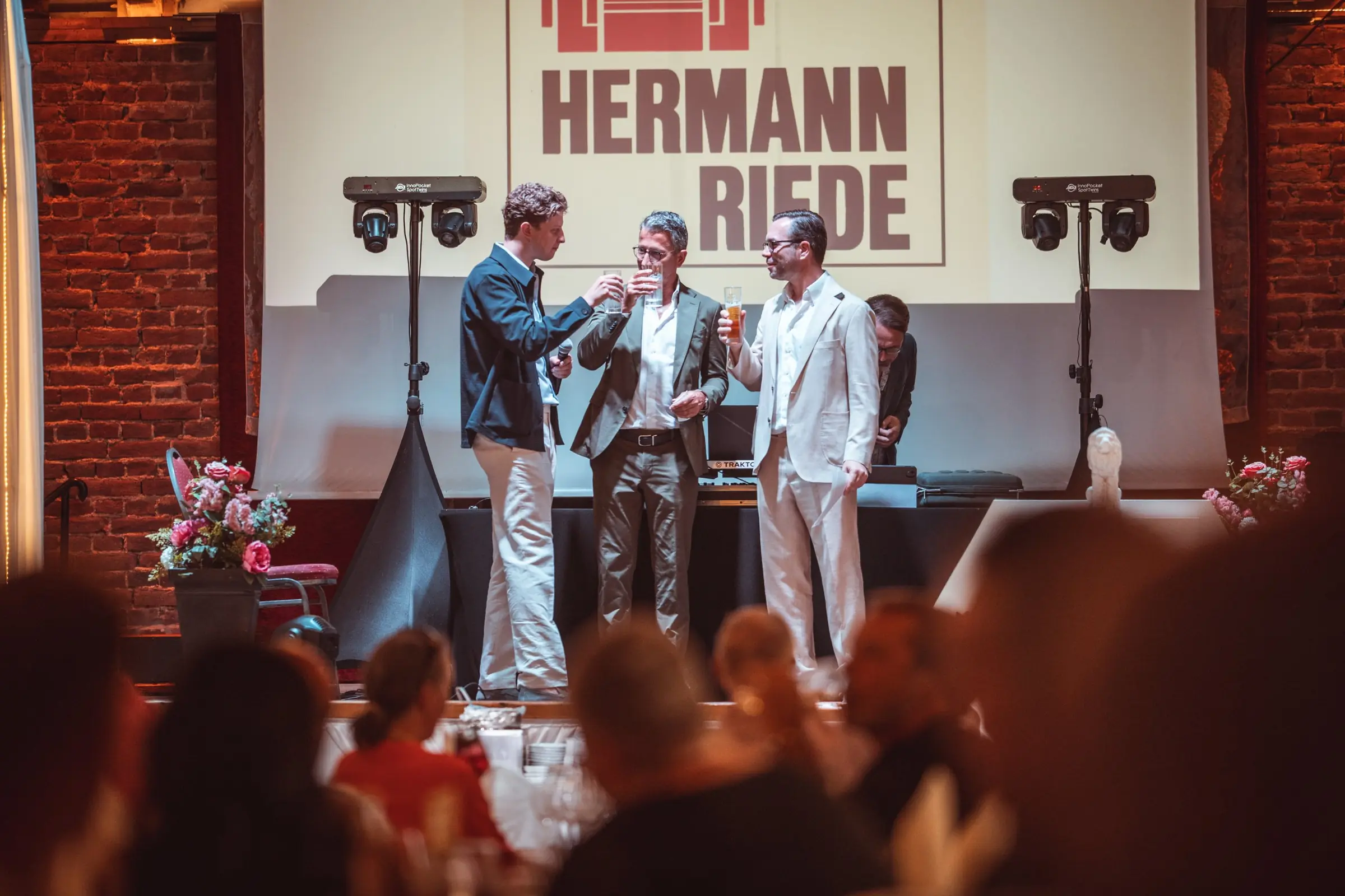 Eventfotografie beim Sommerfest 2026 von HERMANN RIEDE: drei Männer stoßen auf der Bühne vor dem Firmenlogo an